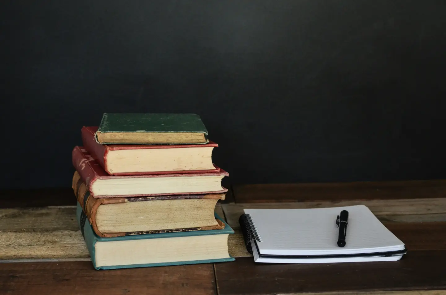 Стопка старых книг и чистый блокнот с ручкой на деревянном столе на фоне тёмной доски