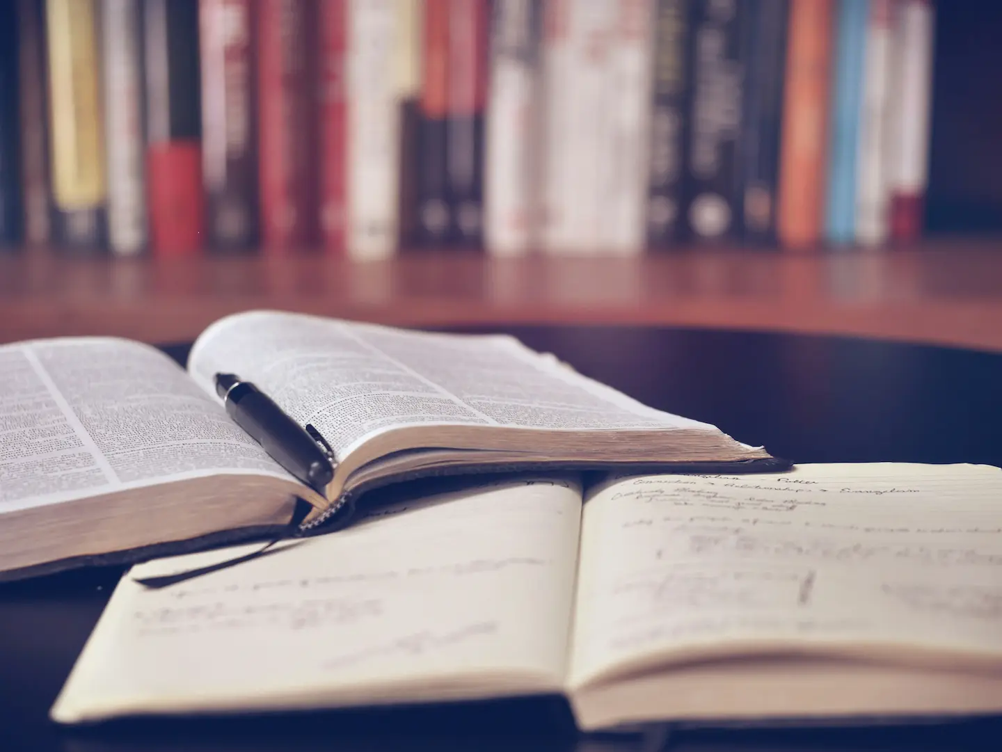 Раскрытая книга с ручкой поверх страниц и блокнот с рукописными заметками на столе, на размытом фоне — полка с книгами