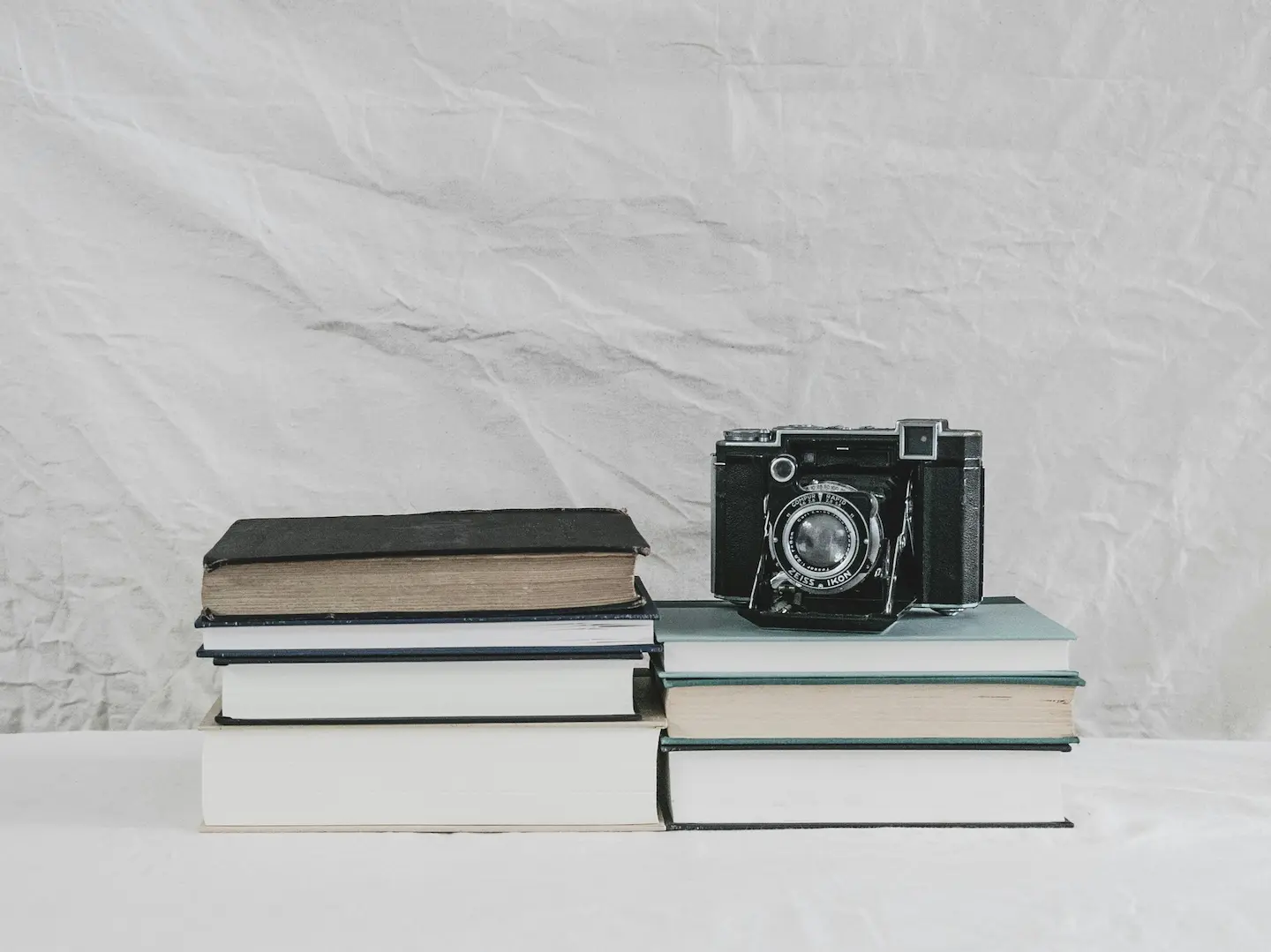 Две стопки книг на светлом фоне, на одной из них стоит старый плёночный фотоаппарат