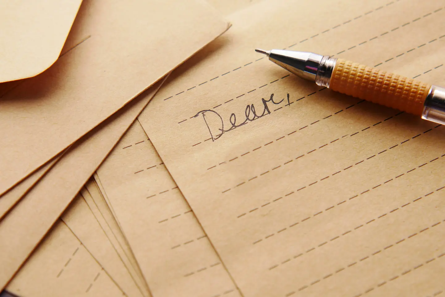 Лист крафтовой бумаги в линейку с первым рукописным словом «Dear,», рядом лежат конверты и ручка